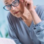 Elderly woman adjusting glasses while reading a book, with text about cataract symptoms, causes, and treatments. Logo for Aarya Eye Care is displayed.