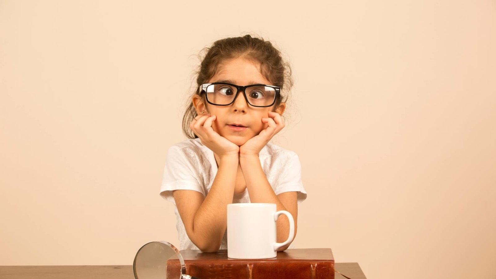 A young girl with oversized glasses rests her chin on her hands, a white mug on the table. Text beside her reads: "Squint: Everything You Should Know About Causes, Symptoms, and Treatment.