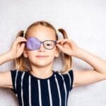 A little girl with glasses smiles while holding a purple eye patch for her lazy eye treatment