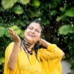 A woman in a yellow dress smiles joyfully while standing in the rain during the monsoon season.