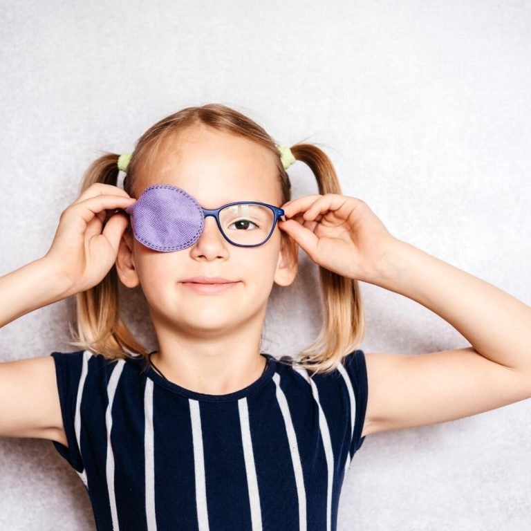 A little girl with glasses smiles while holding a purple eye patch for her lazy eye treatment