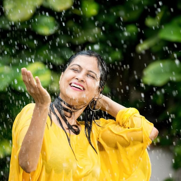 A woman in a yellow dress smiles joyfully while standing in the rain during the monsoon season.
